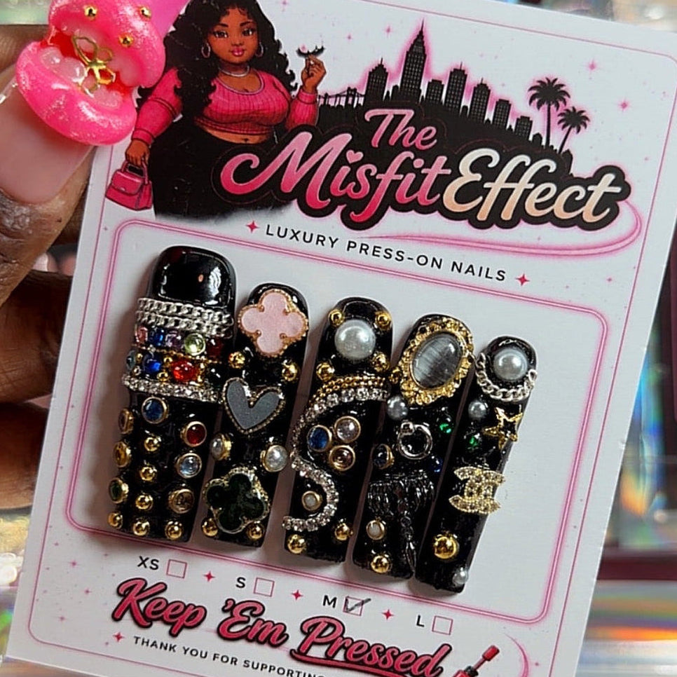 A hand holds a package of 'The Misfit Effect' luxury press-on nails, featuring black coffin-shaped nails heavily embellished with gold studs, rhinestones, and silver heart charms. The background is a busy retail display of other colorful press-on nail packages.