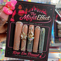 A hand holds a package of 'The Misfit Effect' luxury press-on nails. The package contains five faux nails: three nude beige with gold glitter, one sparkly silver, and one glittery green.