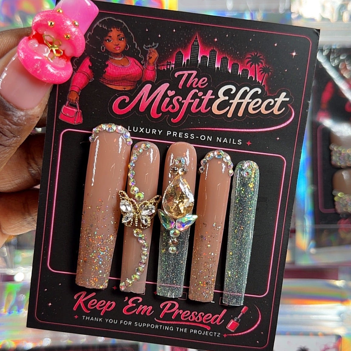A hand holds a package of 'The Misfit Effect' luxury press-on nails. The package contains five faux nails: three nude beige with gold glitter, one sparkly silver, and one glittery green.