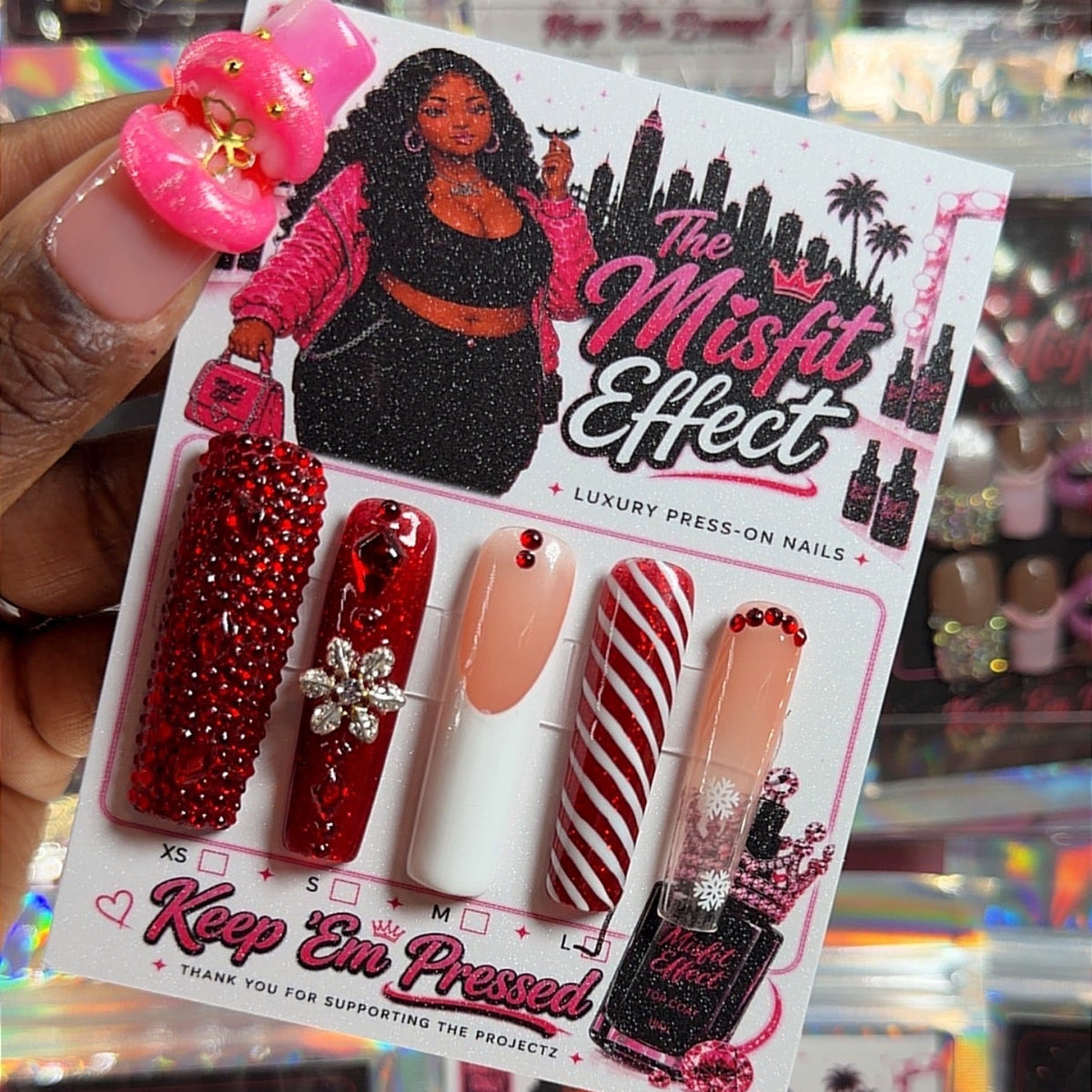 A hand holds a package of 'The Misfit Effect' press-on nails in a retail store setting. The package contains five artificial nails in red glitter, solid white, red-and-white stripes, and nude pink, along with a black press-on nail bottle.