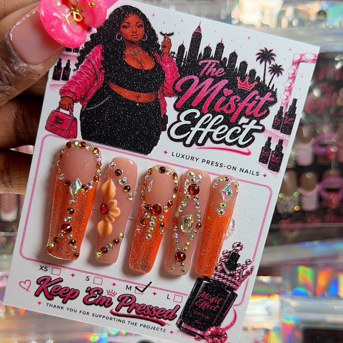 A hand holds a package of 'The Misfit Effect' press-on nails. The nails feature a gradient from peach to orange with gold glitter and rhinestone embellishments.