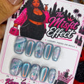 A hand holds a card of press-on nails labeled 'The Misfit Effect' against a busy background of jewelry and accessories. The nails are metallic light blue with silver glitter and a small green gem design.