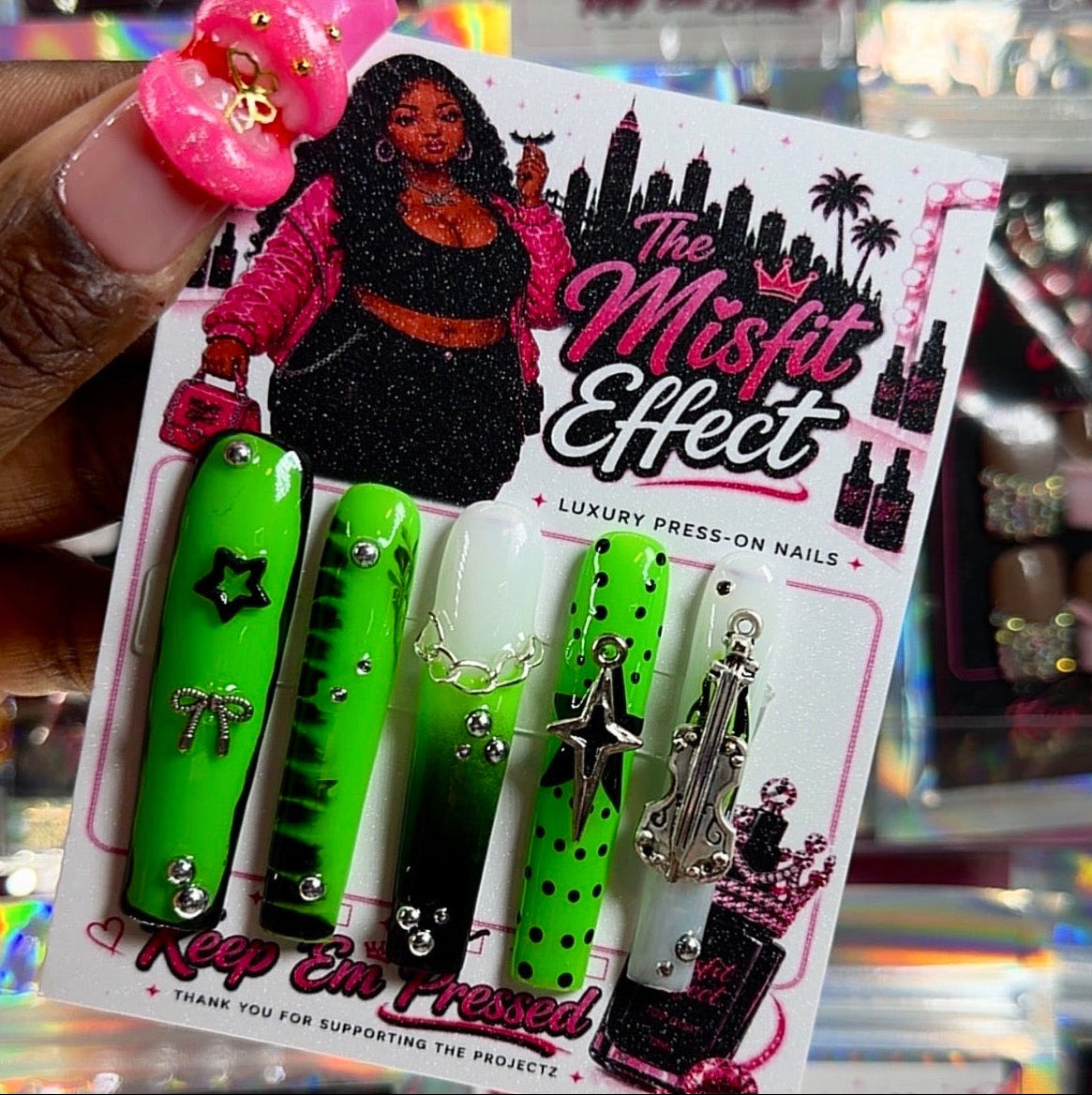 A hand holds a package of 'The Misfit Effect' press-on nails in a retail store setting. The nails are bright neon green, black, and white with silver rhinestone accents.